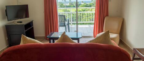 A room with a red sofa, a TV, a small table, and a balcony with a view of the sea.