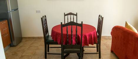 A dining area with a round table covered by a red tablecloth, surrounded by four chairs. A red sofa is positioned to the right of the table.