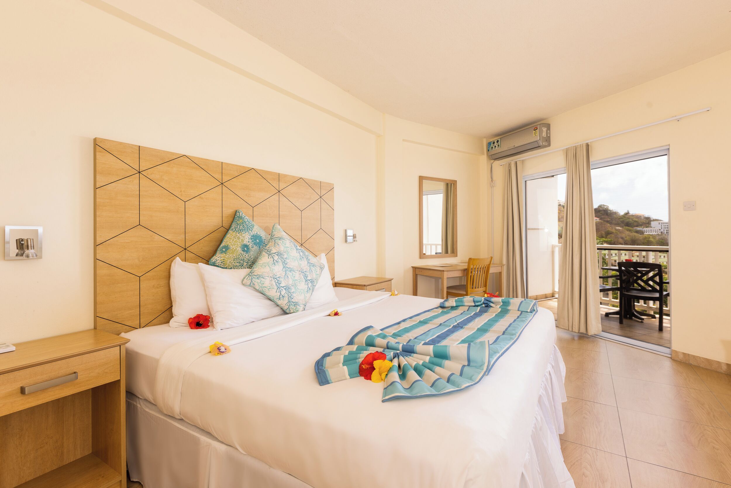 A hotel room with a large bed, a wooden headboard, a bedside table, a chair, a small table, and a balcony with a view.