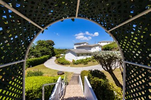 A view through a lattice structure with a pathway leading to a white building surrounded by greenery.