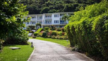 Un hotel con un camino pavimentado que conduce a un edificio blanco con varios balcones, rodeado de exuberante vegetación y un paisaje montañoso.