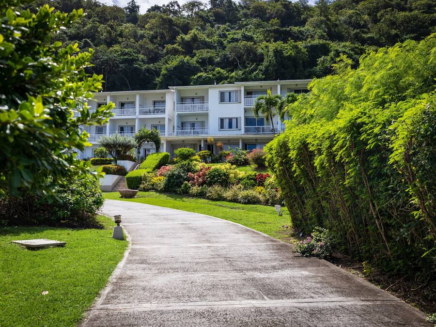 Un hotel con un camino pavimentado que conduce a un edificio blanco con varios balcones, rodeado de exuberante vegetación y un paisaje montañoso.