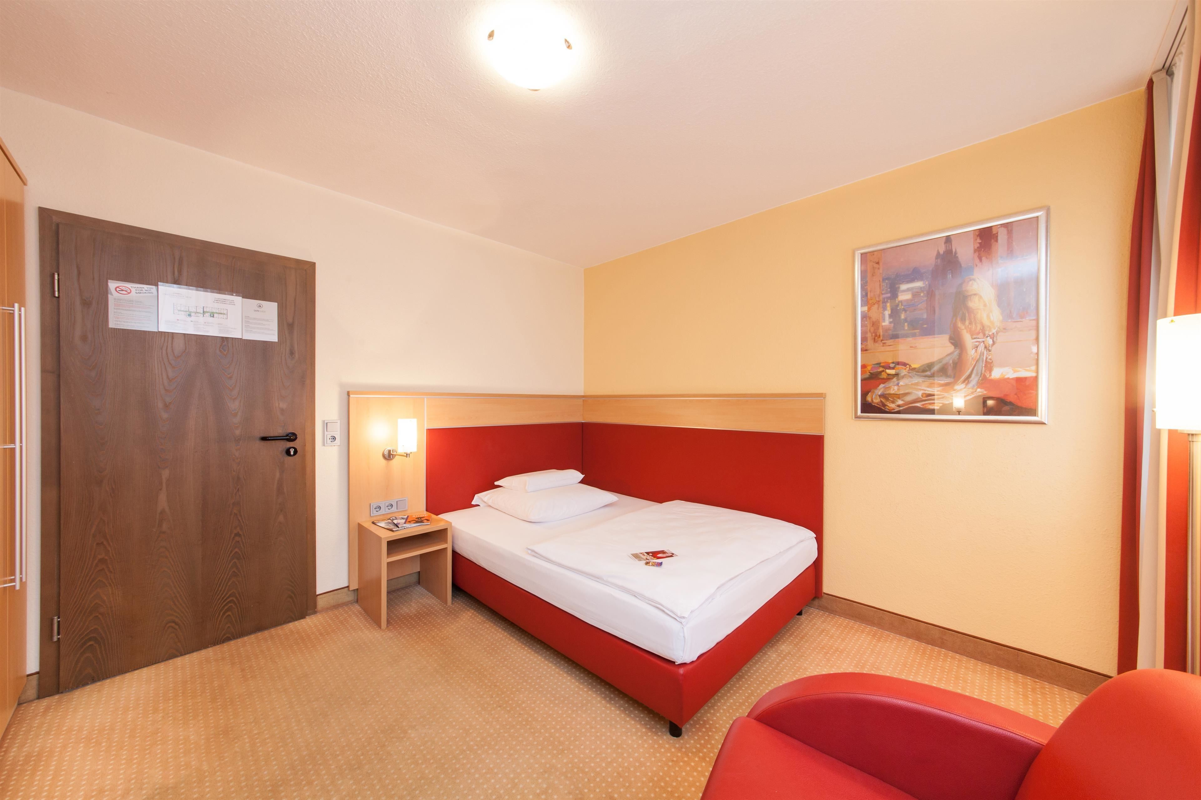 A hotel room with a red bed, a wooden door, a nightstand, a red chair, and a painting on the wall.