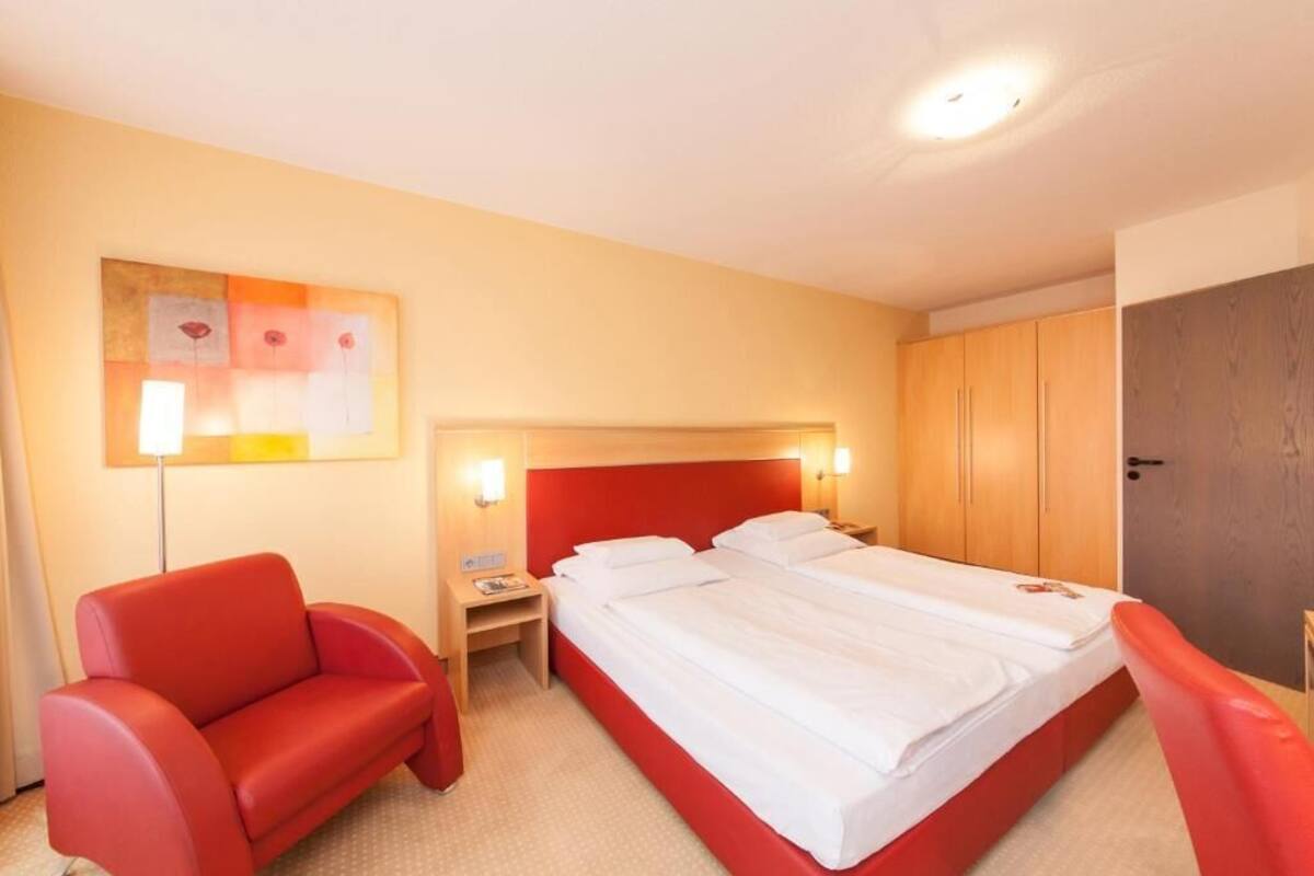 Une chambre d’hôtel avec un grand lit, un fauteuil rouge, une table de chevet avec une lampe et une armoire en bois.