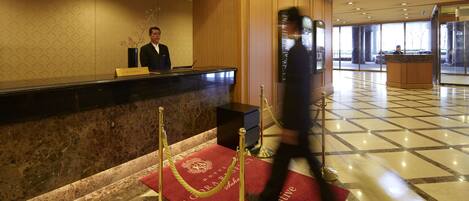 A hotel reception area with a reception desk, a red carpet with "Executive Floor" text, and a security guard walking by.