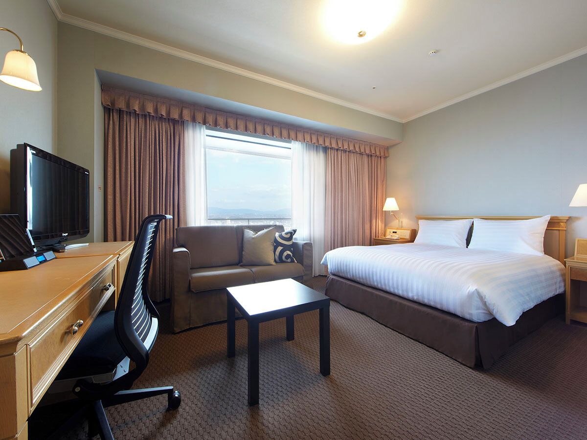 Ein Hotelzimmer mit Bett, Schreibtisch mit Fernseher, Stuhl, Sofa, Tisch und einem Fenster mit Vorhängen.