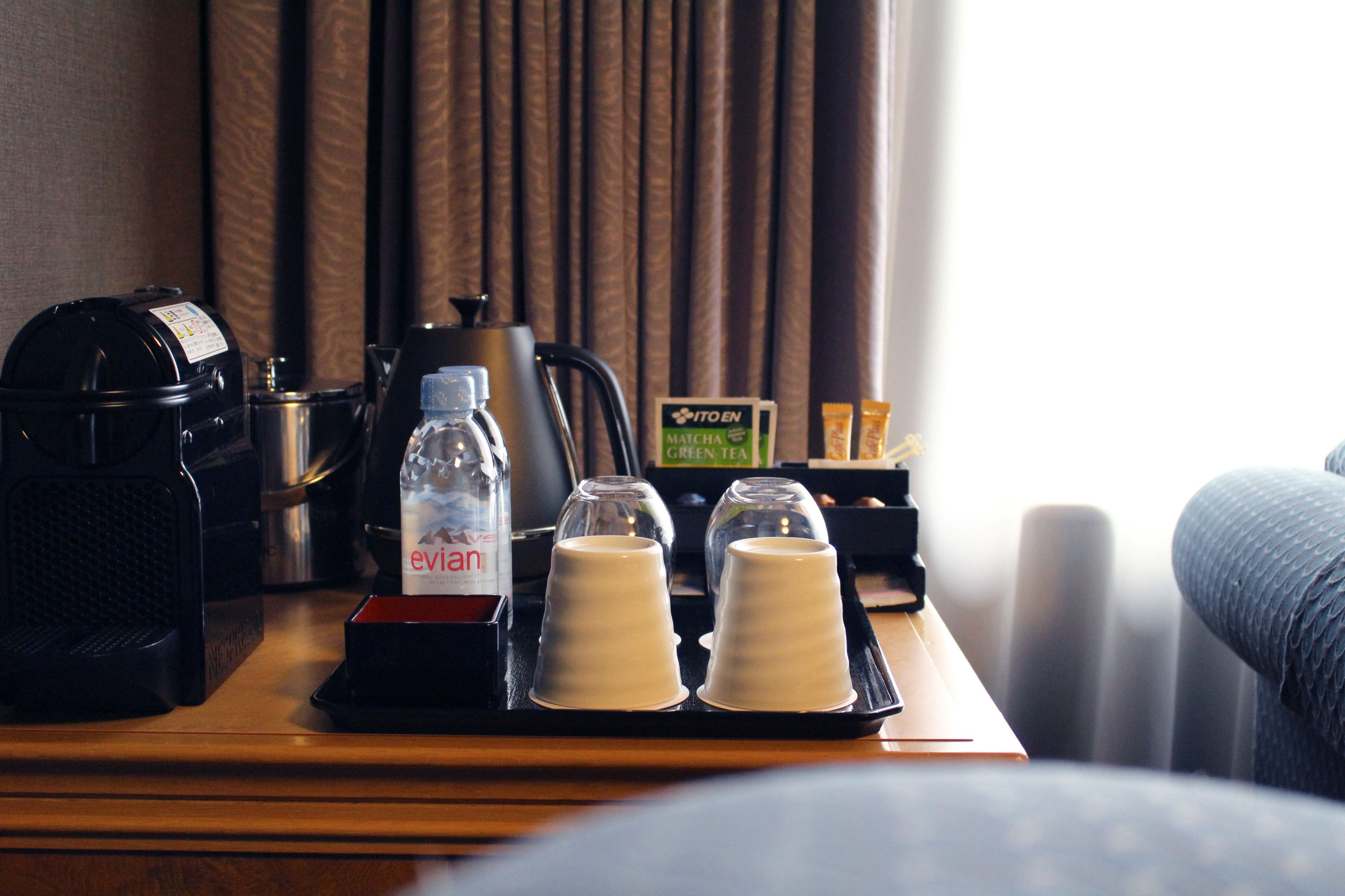 Una habitación de hotel con una cafetera, una botella de Evian, dos vasos y un menú sobre una mesa de madera.