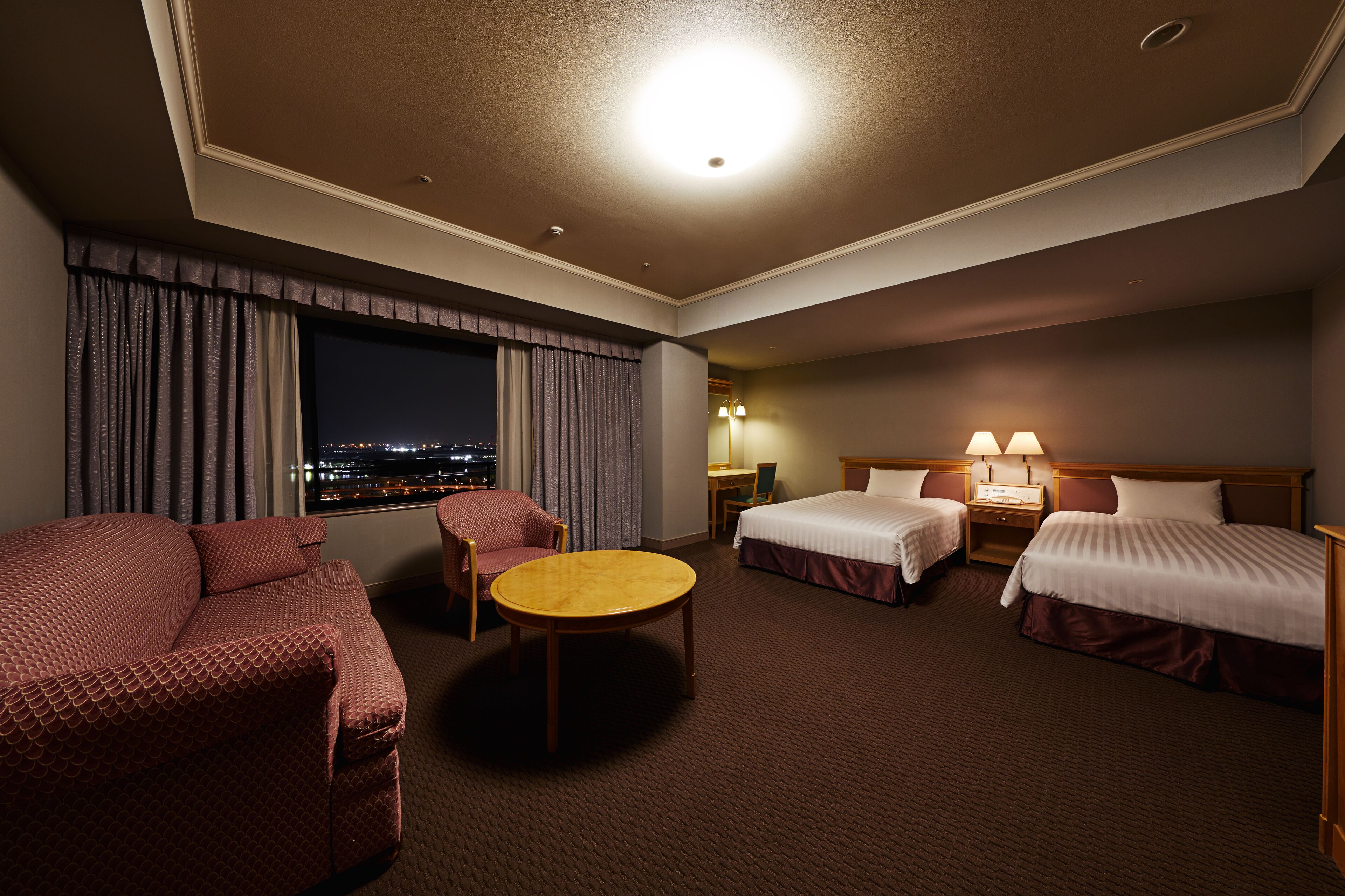 A hotel room with two beds, a sofa, a round table, and a view of the city at night.