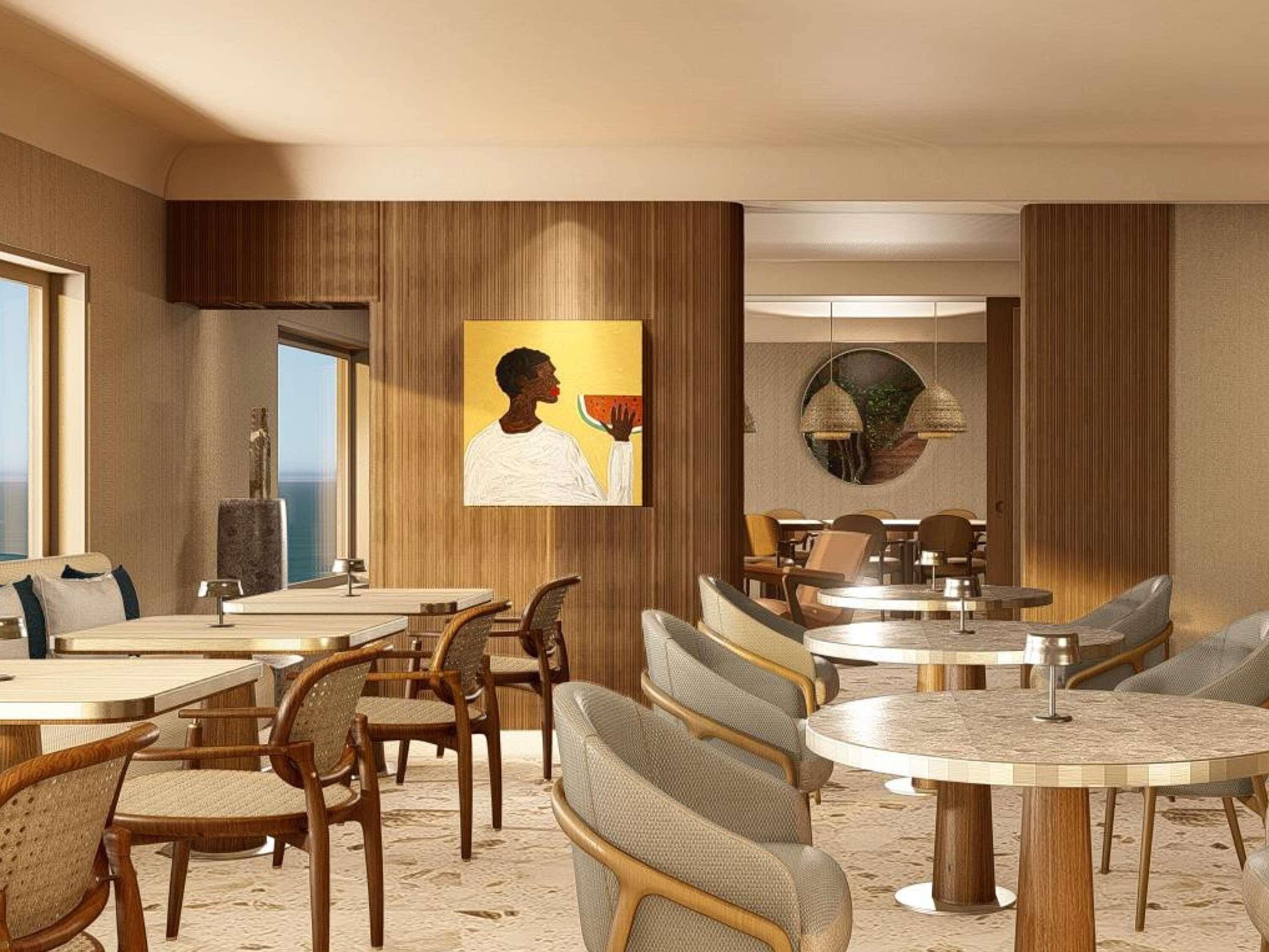 Un comedor moderno con mesas y sillas redondas, un cuadro grande en la pared y vista al mar a través de una ventana.