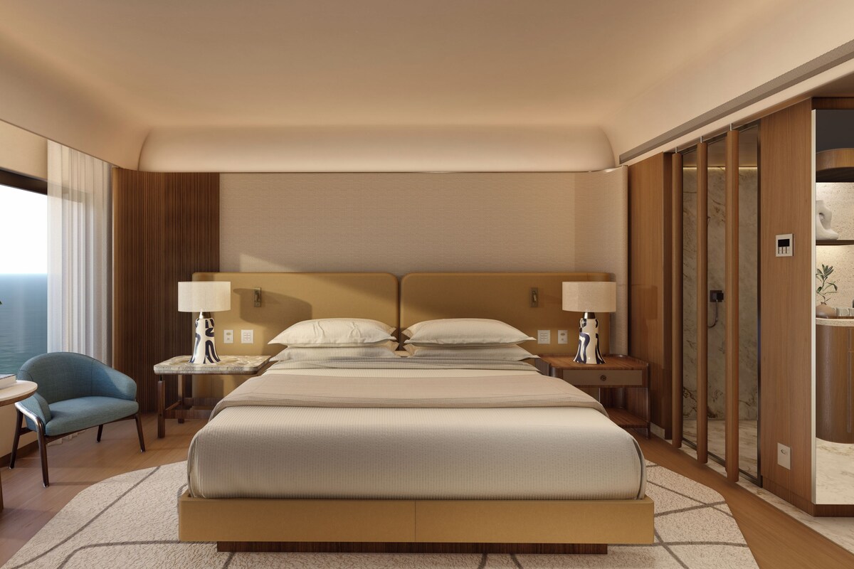 Habitación de hotel con una cama grande, dos mesitas de noche, una silla, una pequeña mesa con una planta y vistas al océano.