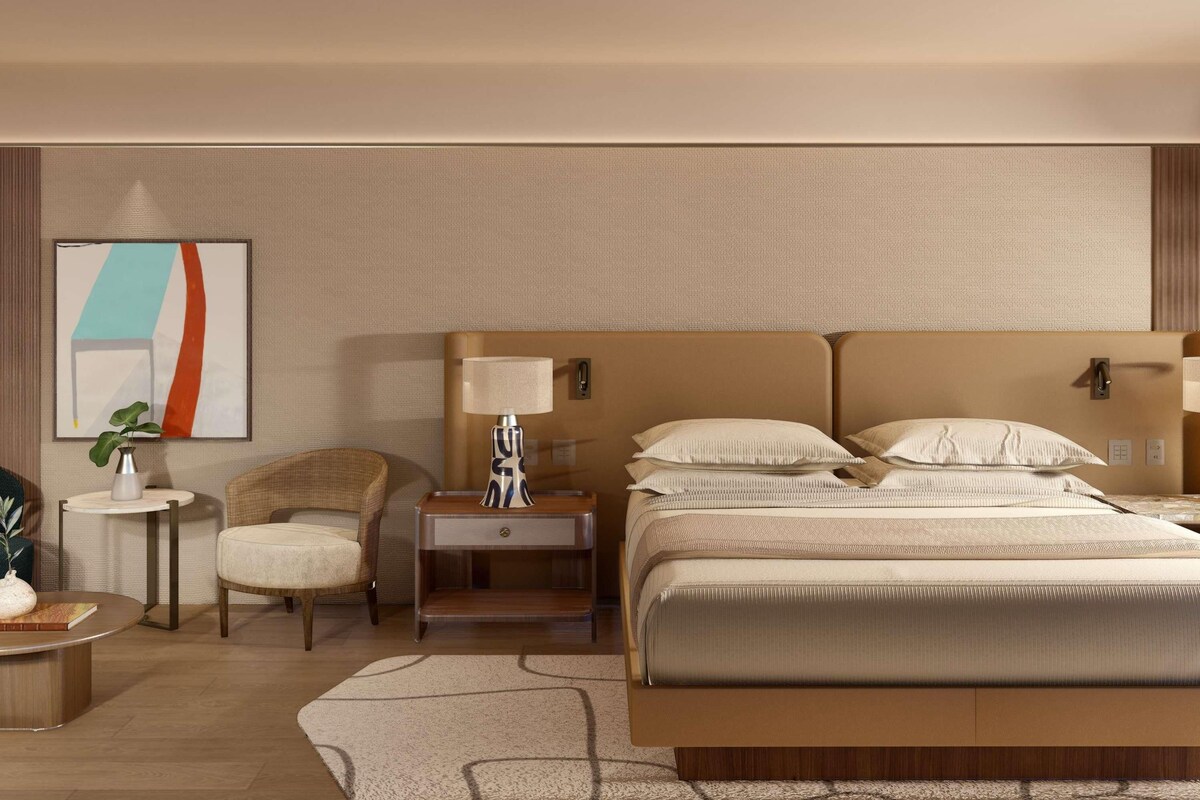 Habitación de hotel con una cama grande, mesitas de noche, una silla, una pequeña mesa con lámpara y una obra de arte en la pared.