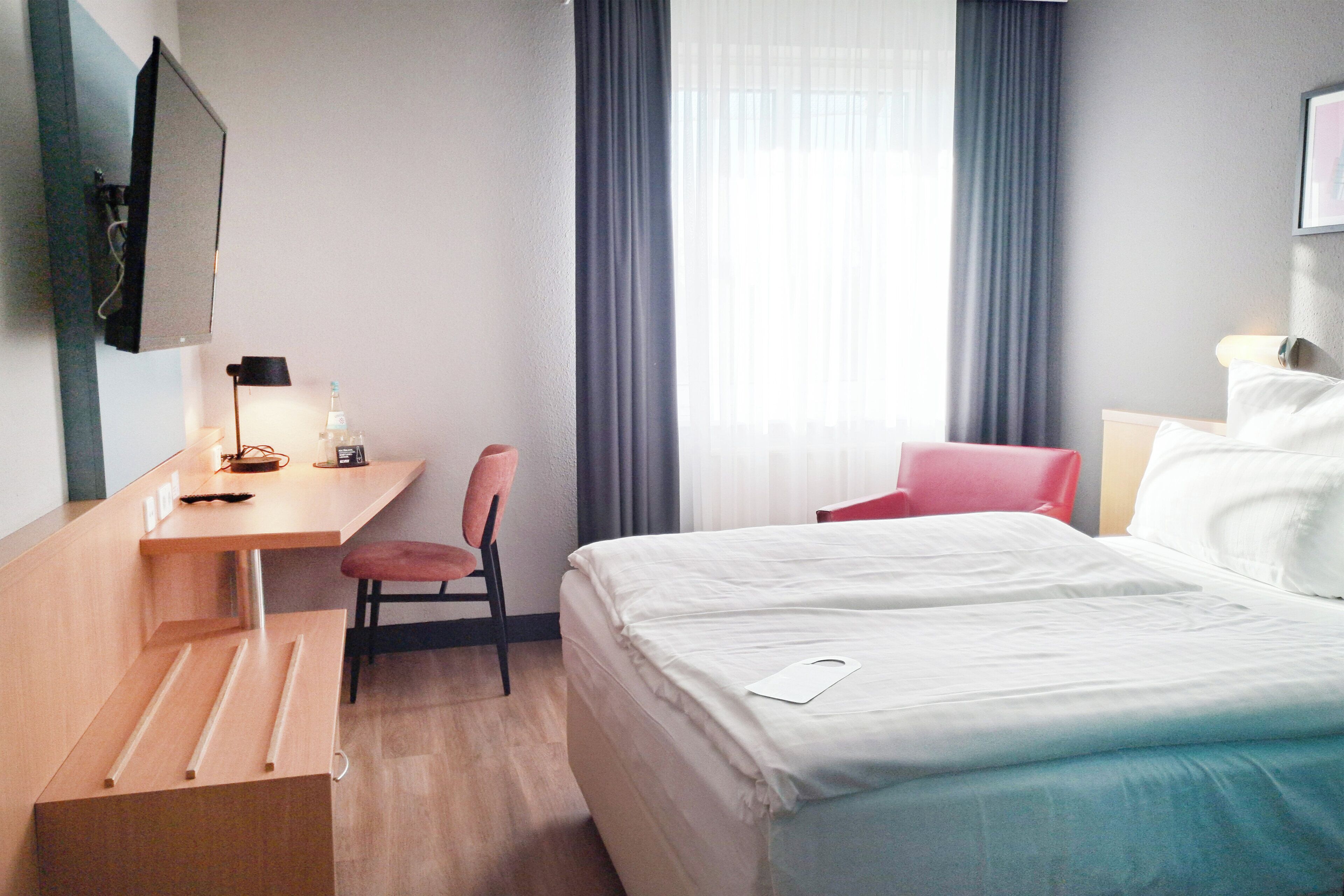A hotel room with a bed, a desk, a chair, a TV, and a window with curtains.