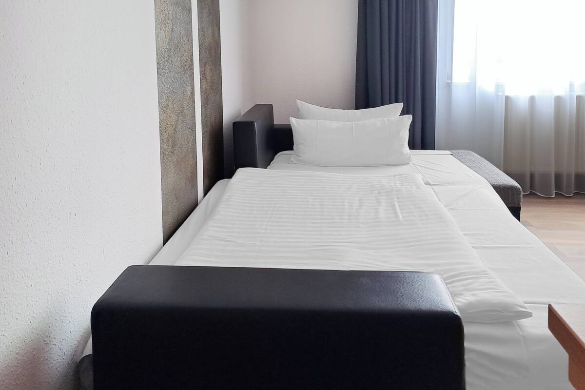 Une chambre d’hôtel avec un lit, un banc noir, une table de chevet et une fenêtre avec des rideaux.