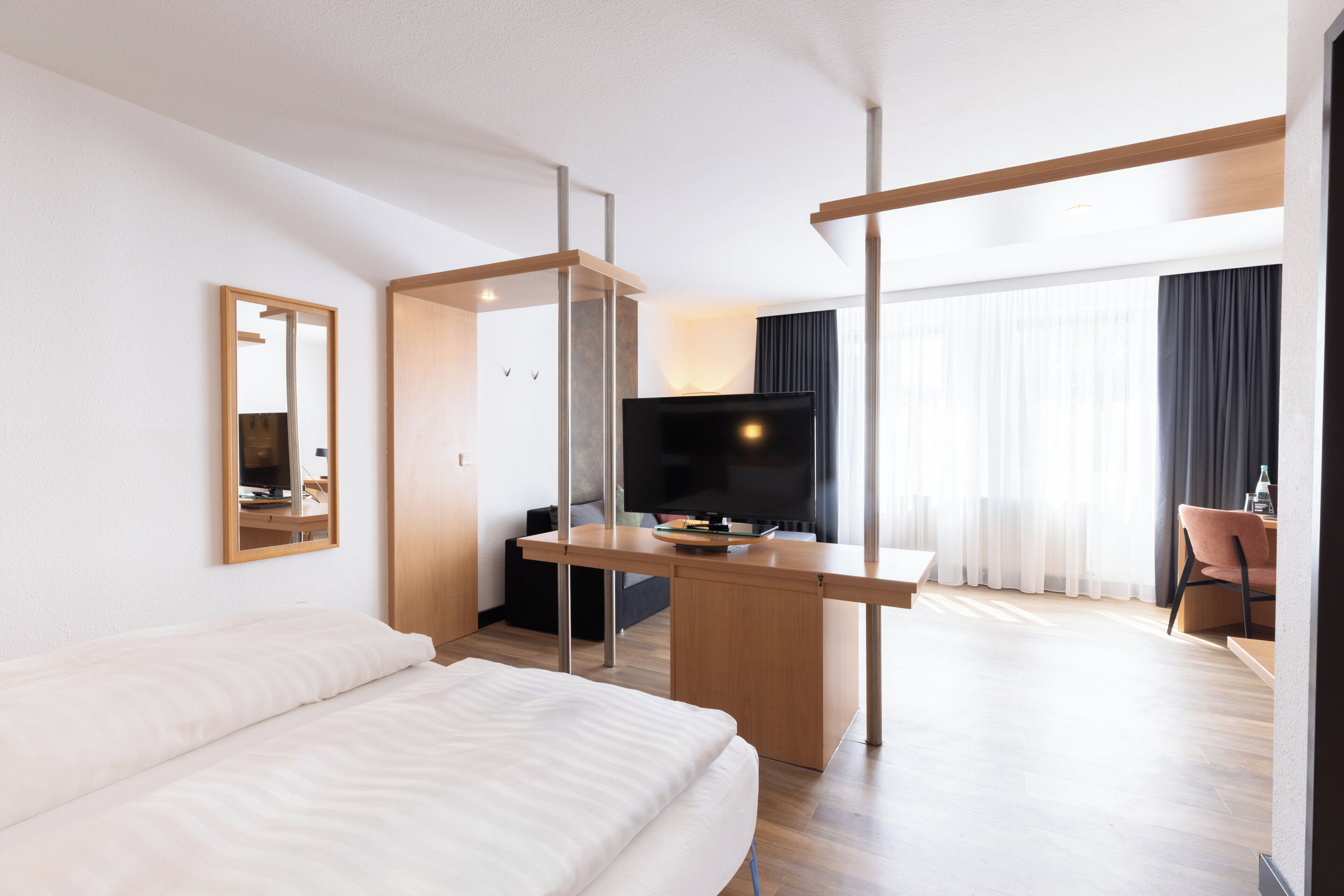 Une chambre d’hôtel avec un lit, un bureau avec une télévision, une chaise et une grande fenêtre avec des rideaux.