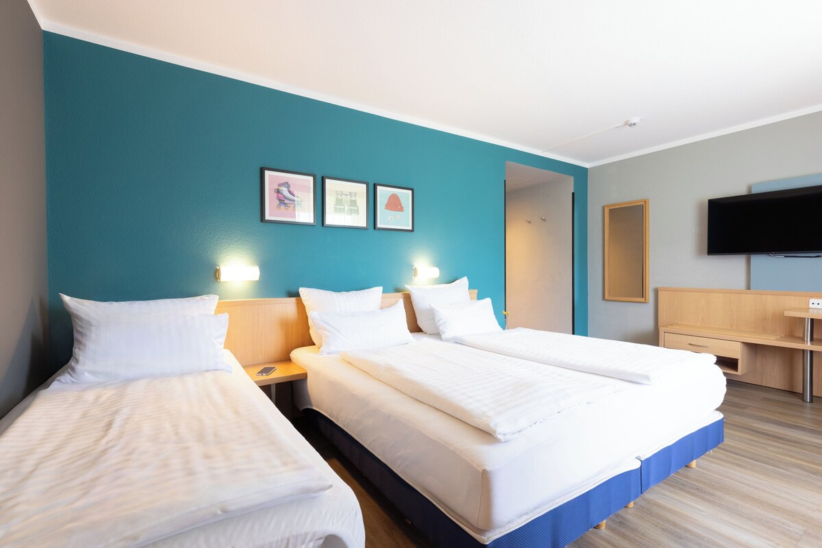 Habitación de hotel con dos camas, un escritorio, un televisor y un baño visible a través de una puerta abierta.