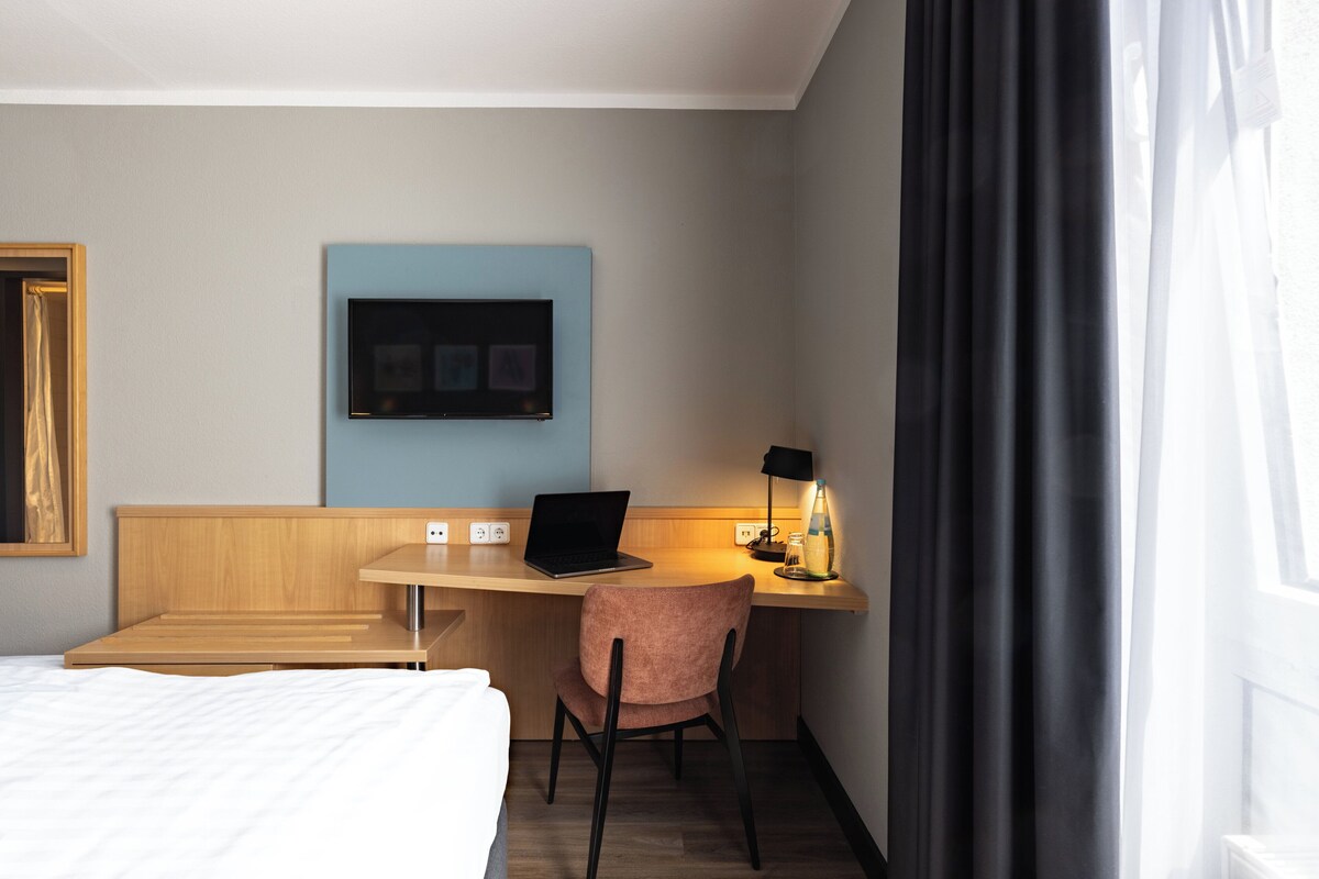 Habitación de hotel con cama, escritorio con computadora portátil, silla, televisor montado en la pared y ventana con cortinas.