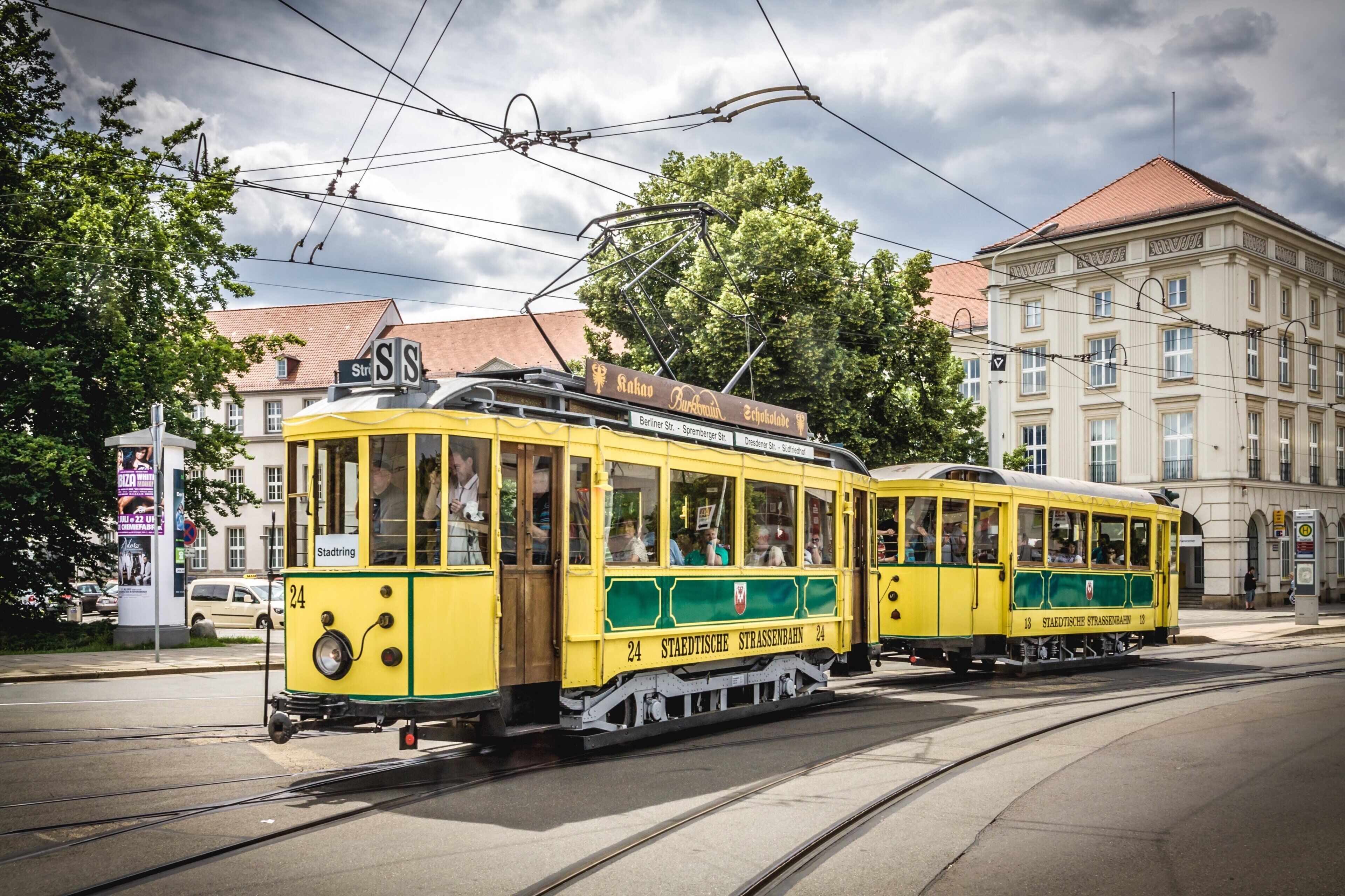 A yellow tram with green accents on a city street, passing by buildings and trees.