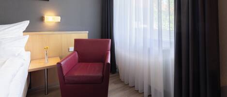 Habitación de hotel con cama, una silla roja, una mesita con un florero, una lámpara de pared y cortinas traslúcidas.