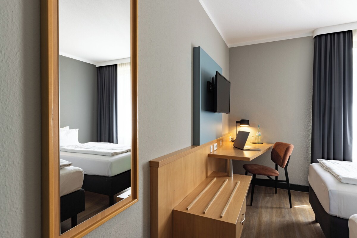 A hotel room with a bed, a desk with a laptop, a chair, a TV mounted on the wall, and a mirror.