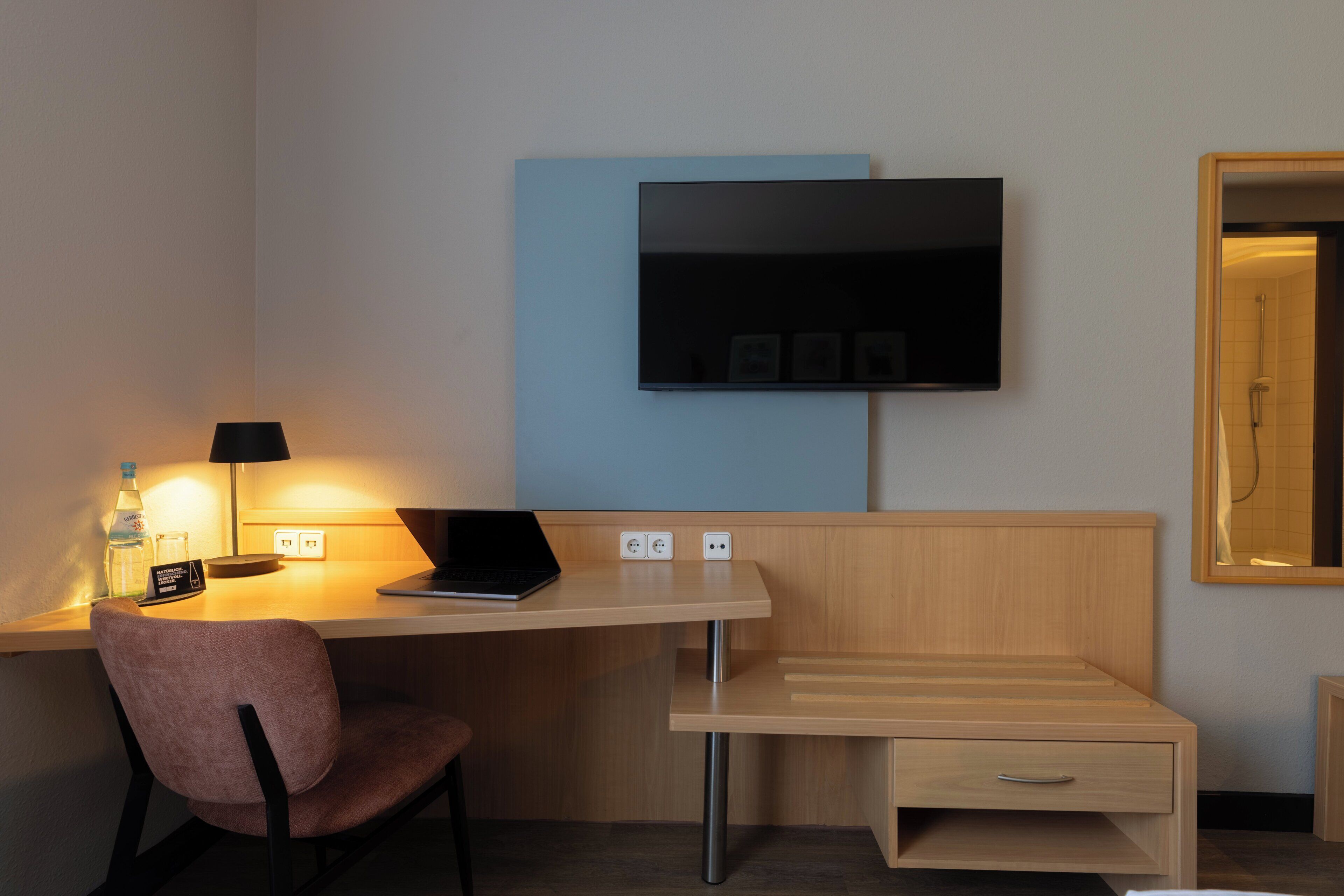 A hotel room with a desk, chair, TV, and a small bedside table.