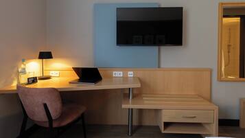 A hotel room with a desk, chair, TV, and a small bedside table.
