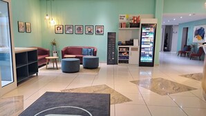 Et hotel lobby med en receptionsdisk, et siddeområde med en rød sofa og skamler, et køleskab til drikkevarer og en væg med indrammede kunstværker.