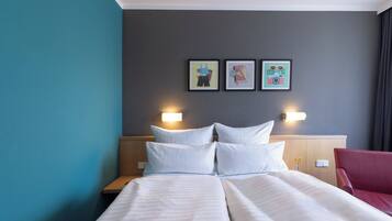 Een hotelkamer met een bed, twee nachtkastjes, een rode stoel en drie ingekaderde foto’s aan de muur.