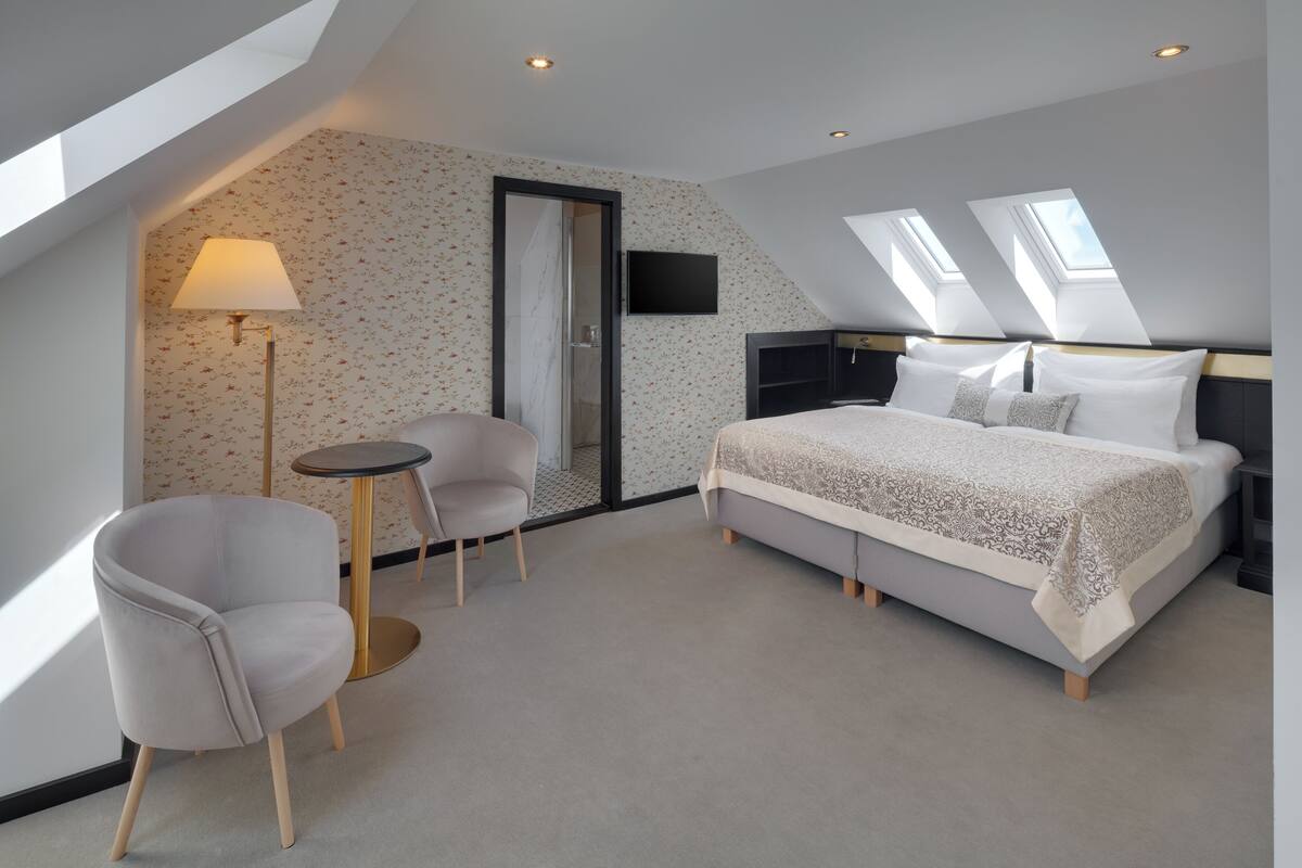 A bedroom with a bed, a chair, a small table, and a TV on the wall.