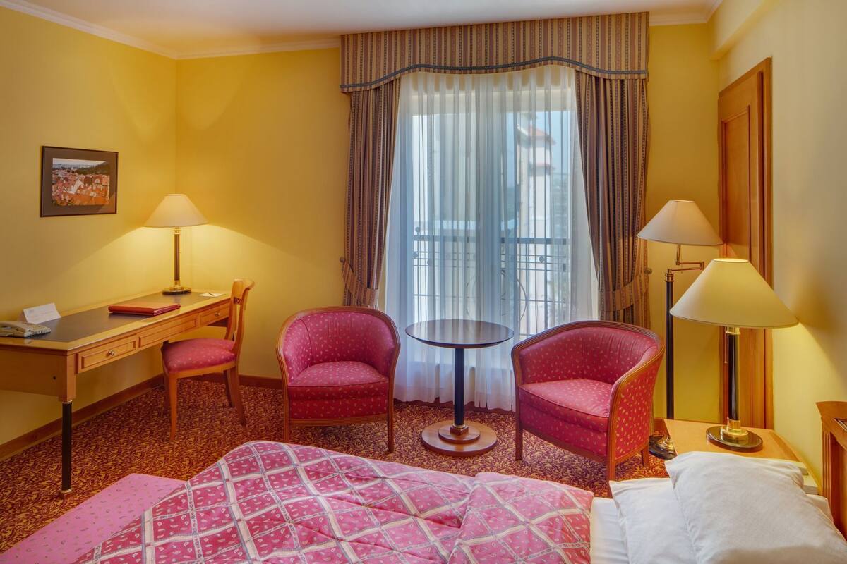 A hotel room with a bed, two armchairs, a desk, a chair, a small round table, and a window with curtains.