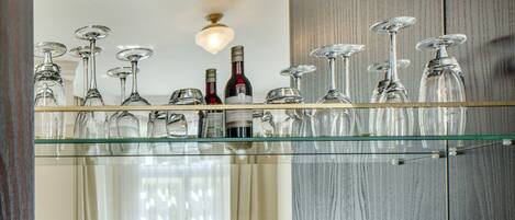 A modern hotel room with a glass shelf displaying wine glasses and bottles, a coffee maker, and a dining area with chairs and a table.