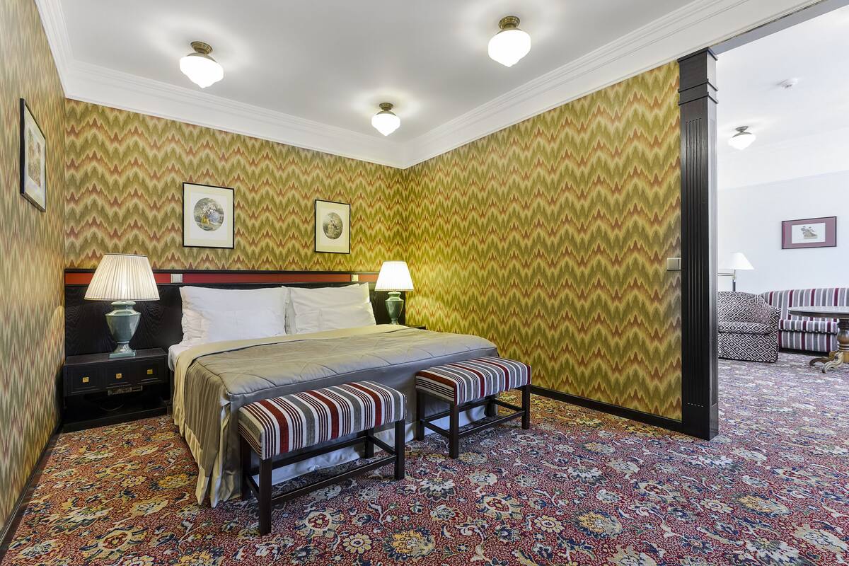 A hotel room with a bed, two bedside tables, a bench, patterned wallpaper, and a carpet.