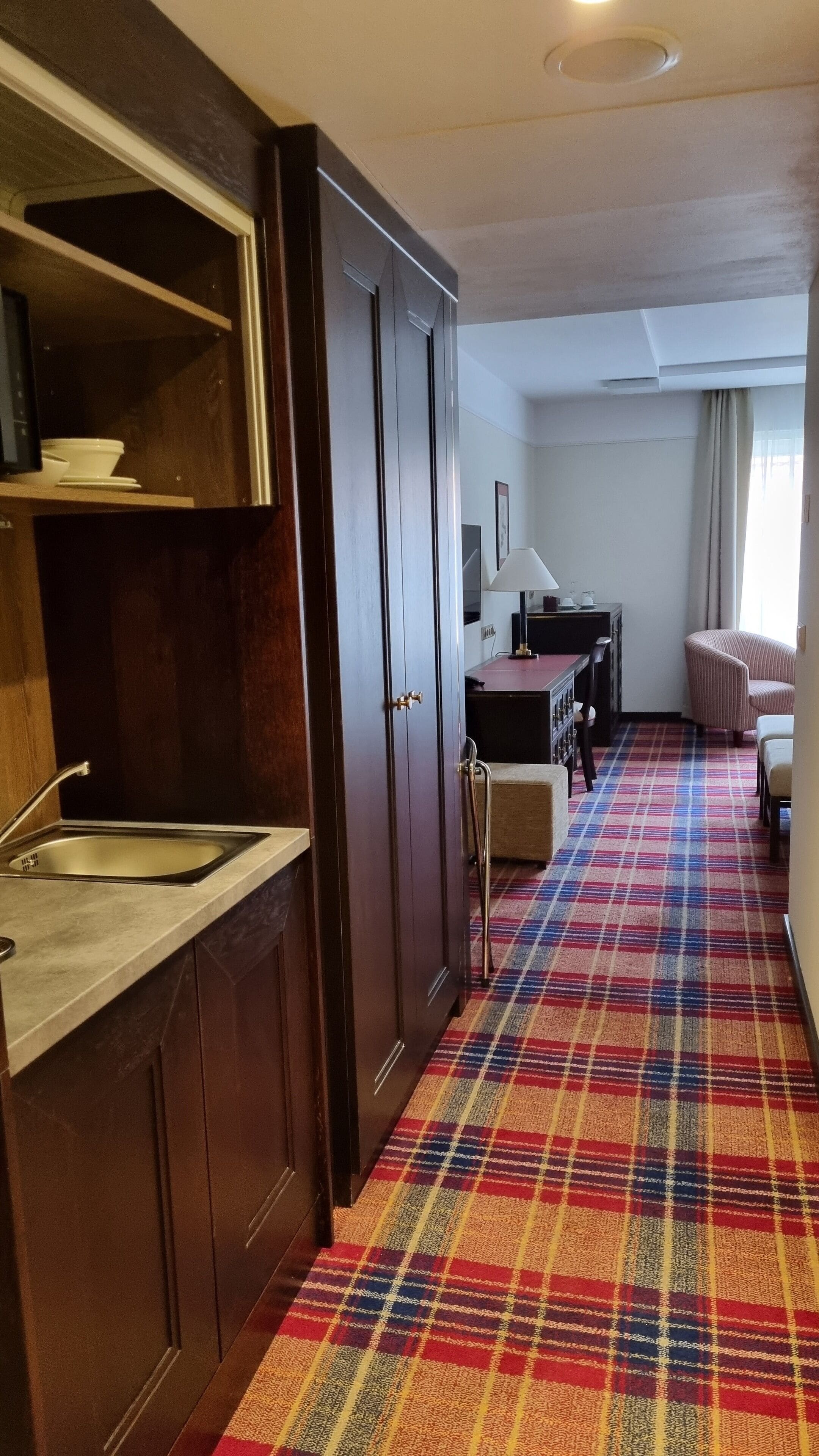 A hotel room with a kitchenette, a plaid carpet, a bed, and a window with curtains.