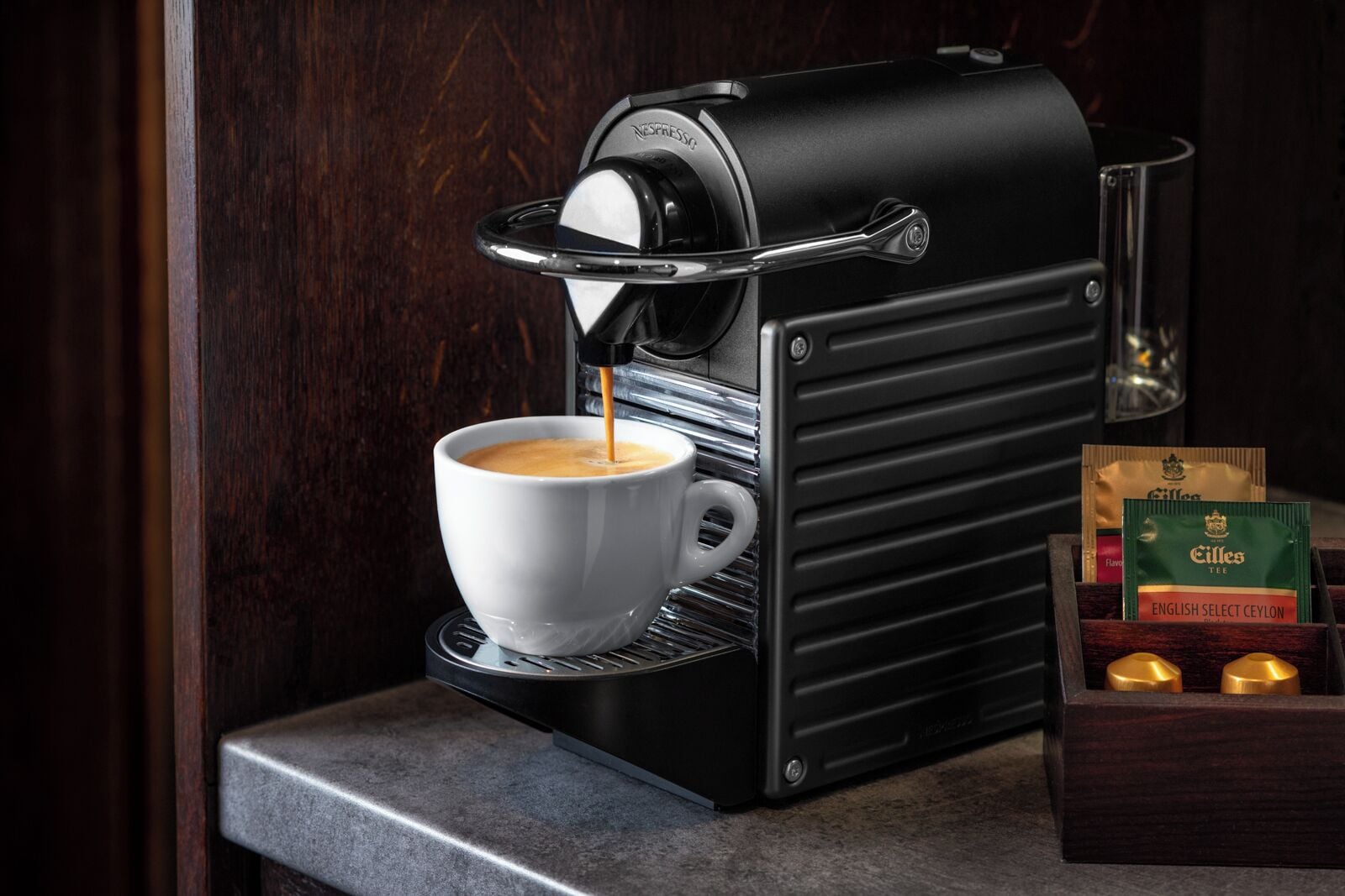 A Nespresso coffee machine dispensing coffee into a cup, with a box of Ceylon tea and two gold-colored items beside it.