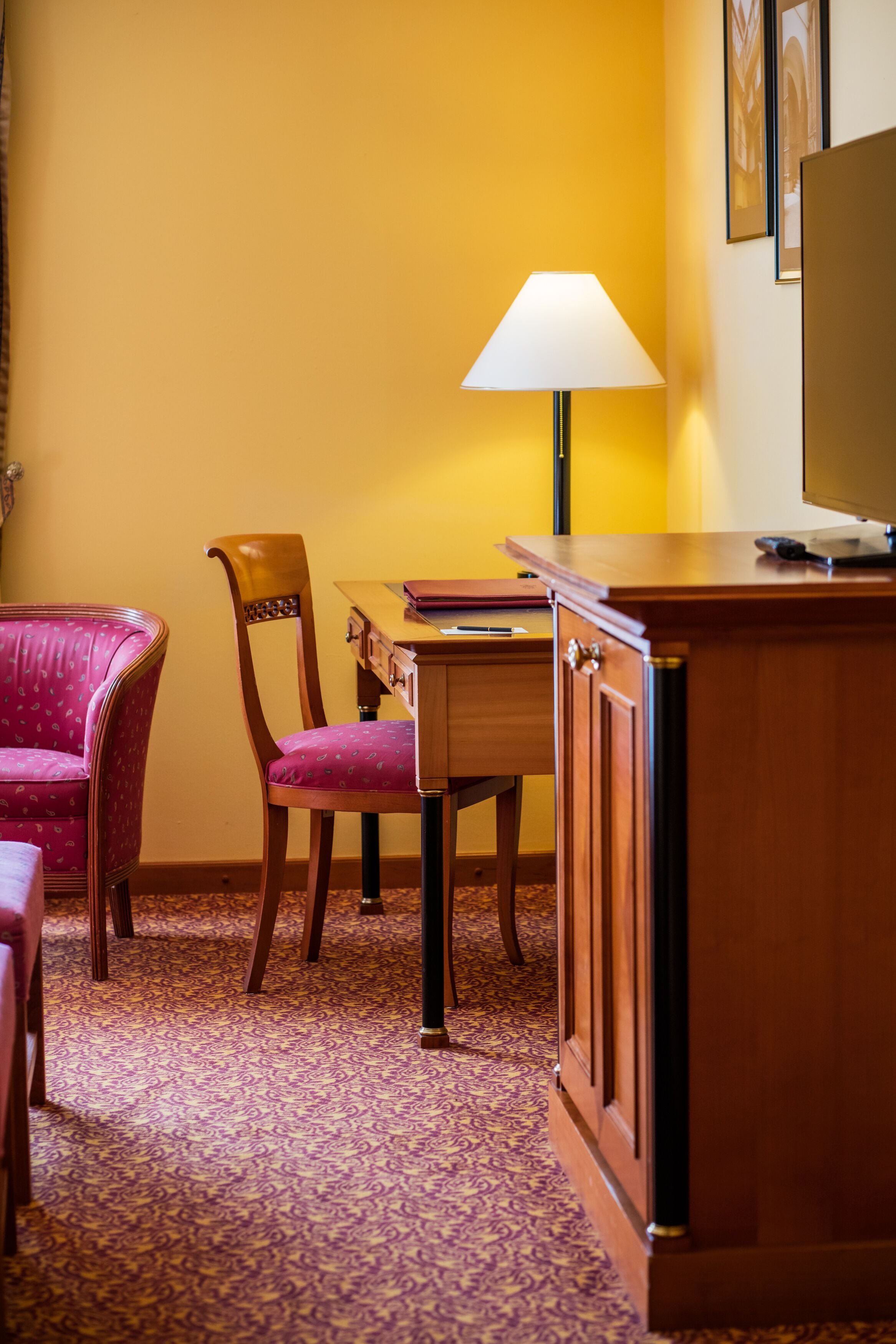 A hotel room with a wooden desk, a chair, a lamp, and a television.