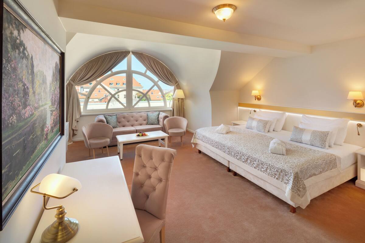 A hotel room with a large bed, a sofa, a chair, a desk, and a large window with a view of buildings.