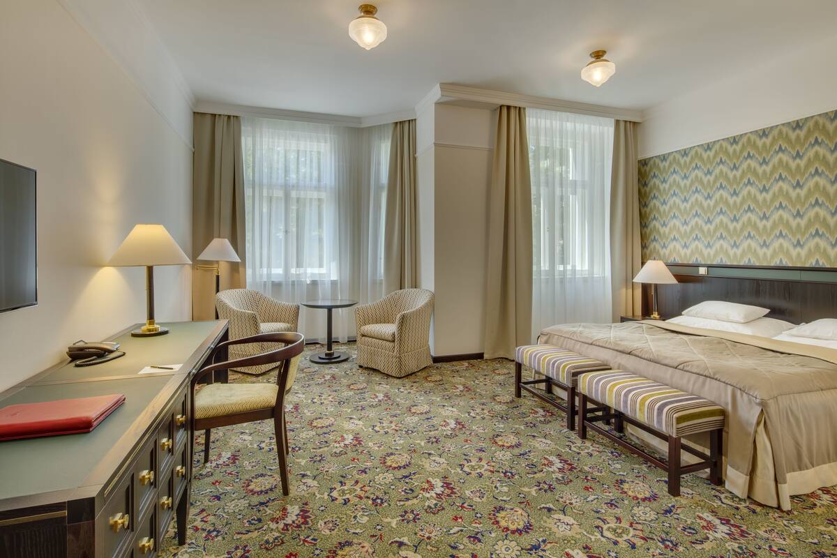 A hotel room with a bed, two chairs, a desk with a lamp, a small table, and a bench.