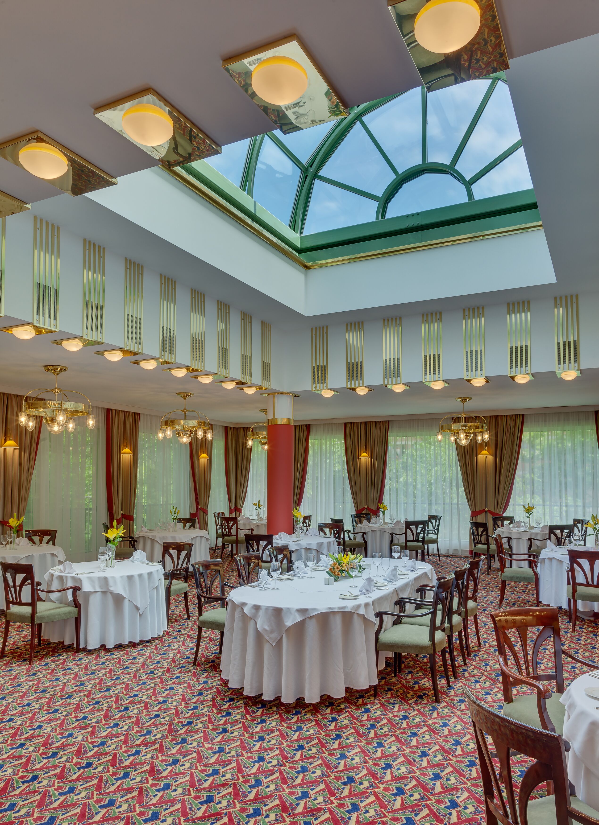 A spacious dining area with round tables, chandeliers, and a high ceiling with skylights.