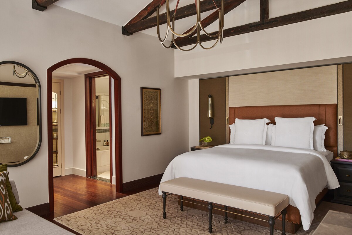 A bedroom with a large bed, a bench, a mirror, and a door leading to another room.