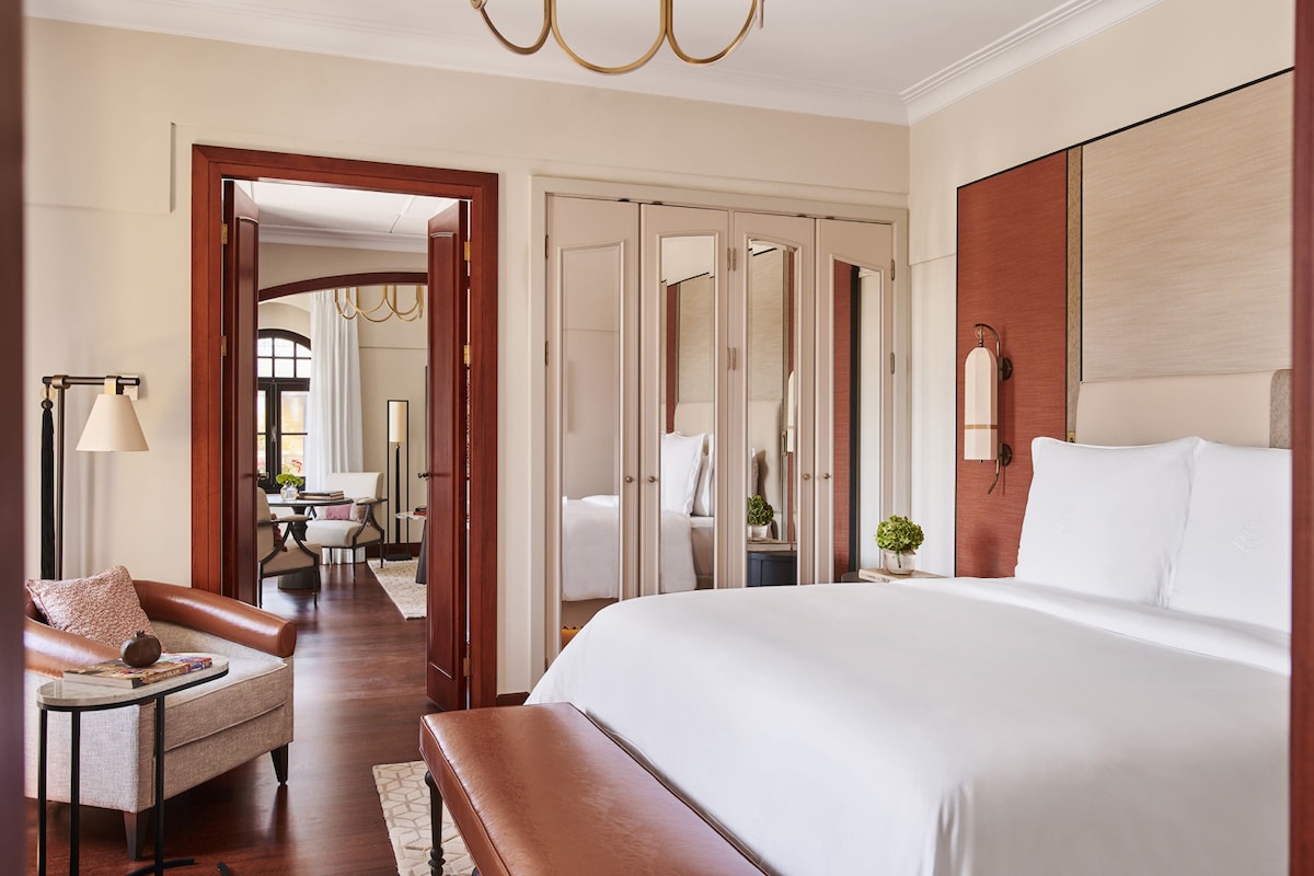 A hotel room with a large bed, a seating area with a bench and armchair, a desk with a lamp, and a wardrobe with mirrors.