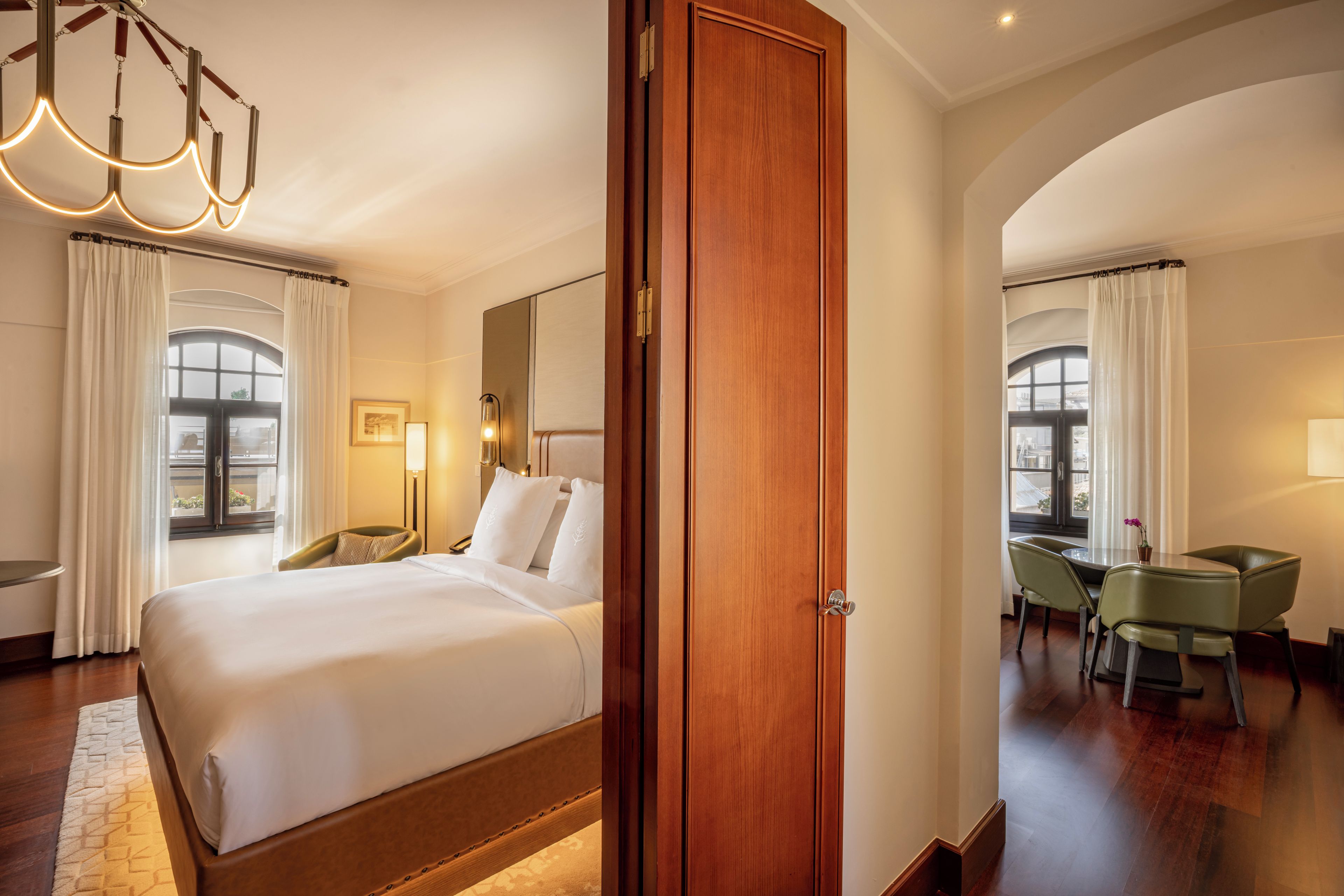 A hotel room with a large bed, a desk, and a chair, a chandelier, and a view through the window.