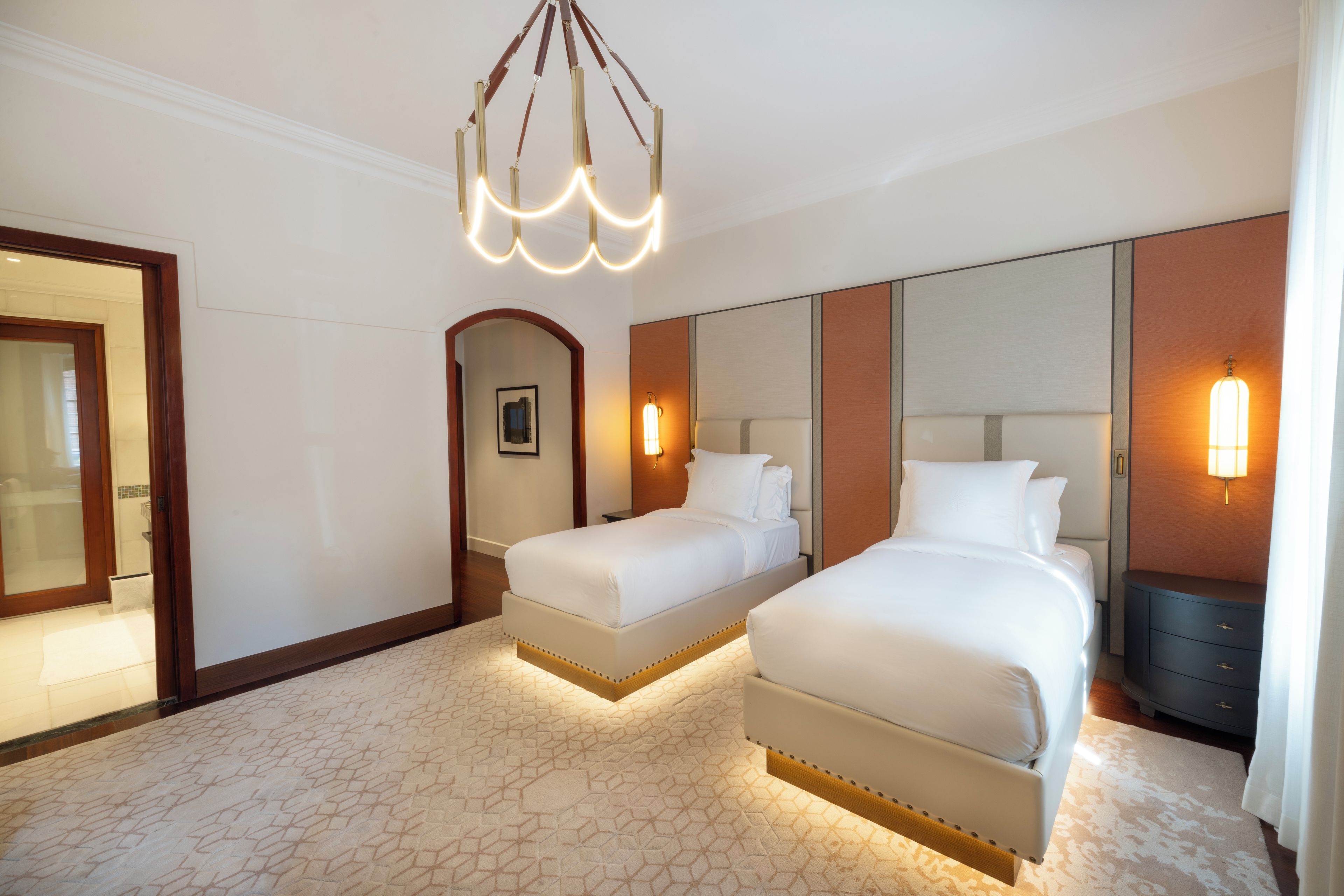 A hotel room with two beds, a large mirror, a chandelier, and a nightstand with a lamp.