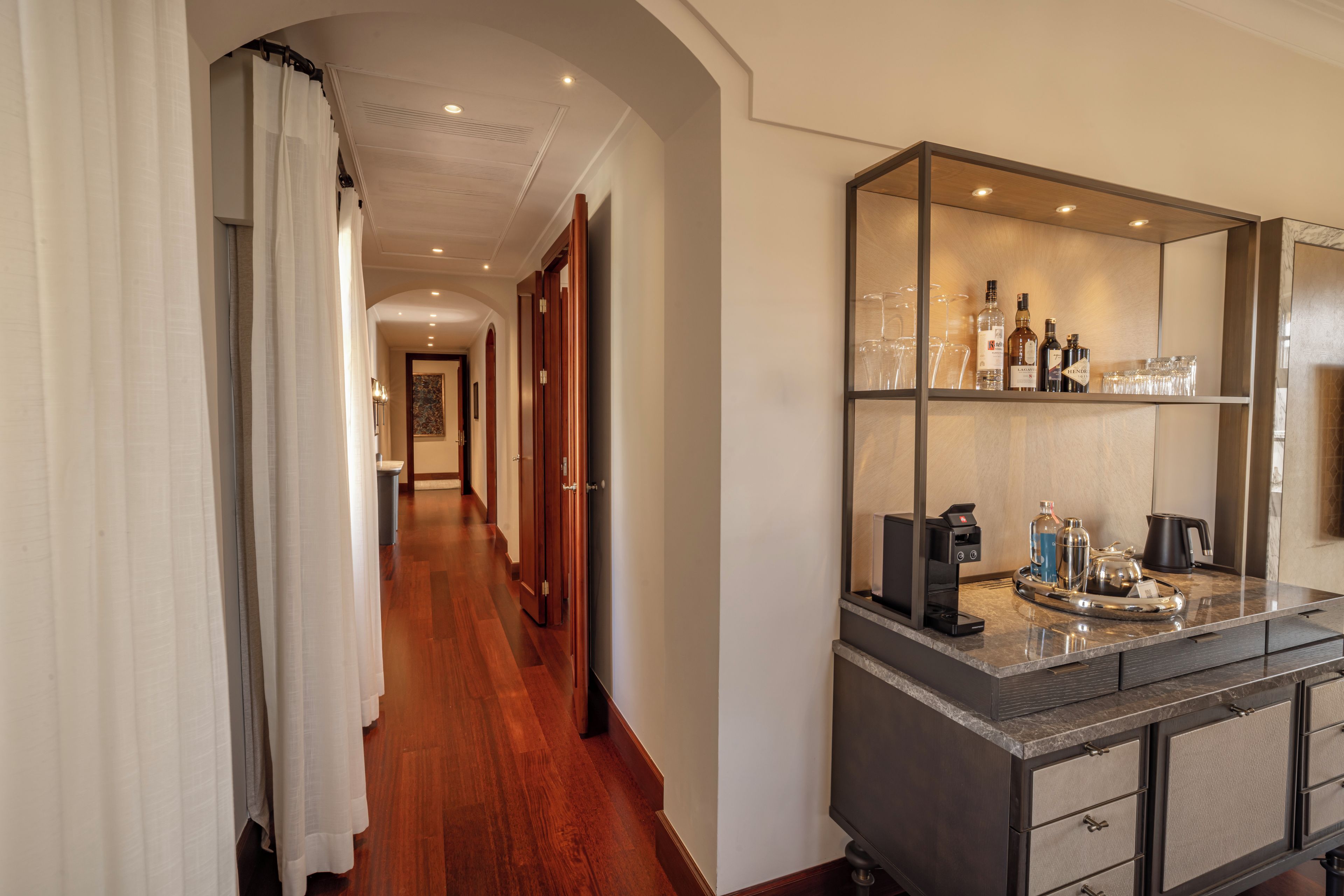 Un corridoio d'hotel con pavimento in legno, un grande specchio e un minibar con diverse bottiglie e bicchieri.