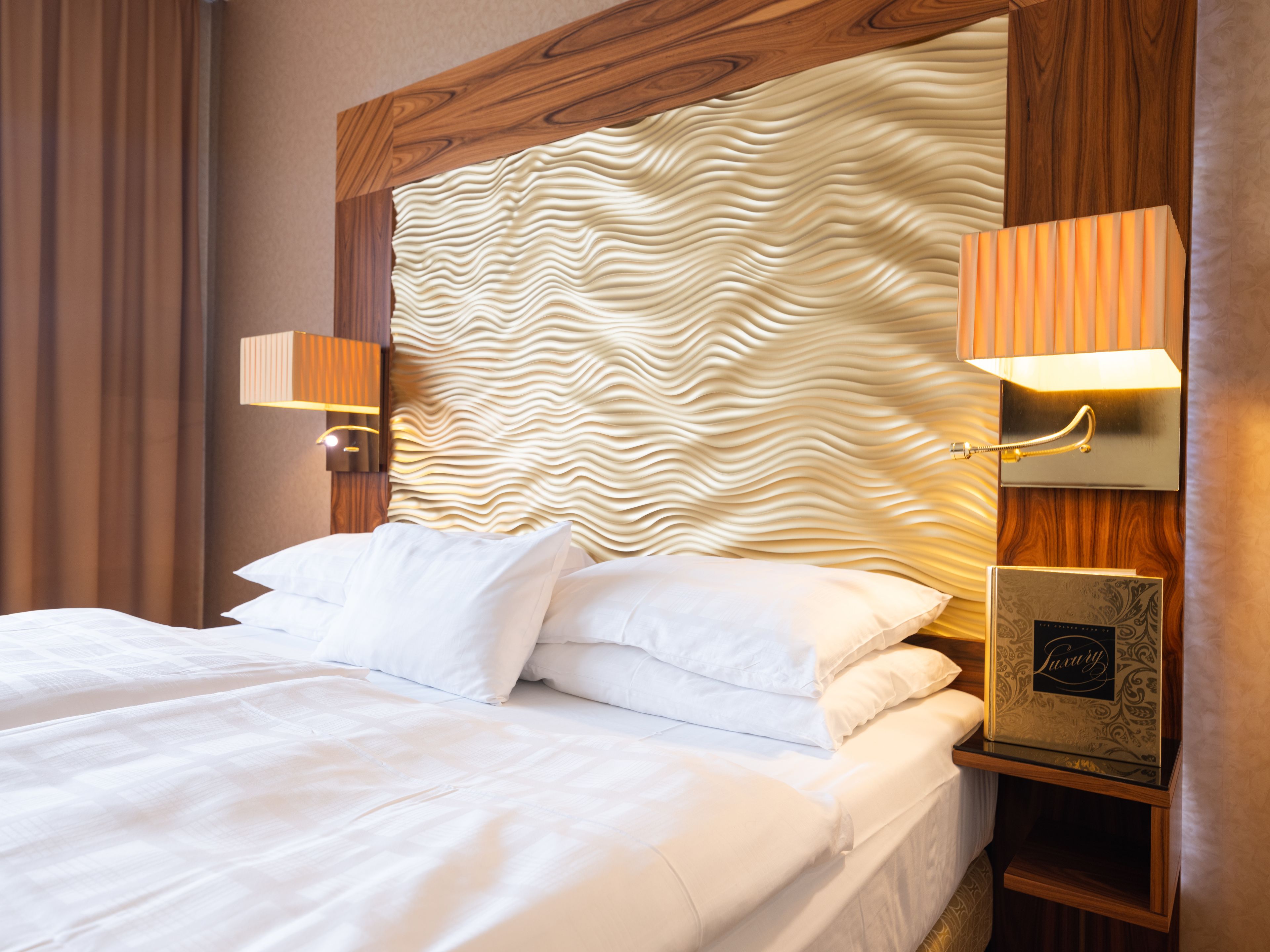 A neatly made bed with white linens, a wooden headboard with a wavy pattern, and two bedside lamps on either side.