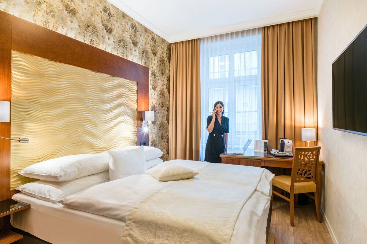 Quarto de hotel com uma cama grande, uma escrivaninha com um laptop, uma cadeira e uma janela ampla com cortinas.