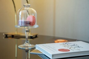 Μια γυάλινη θολωτή προθήκη με μακαρόν τοποθετημένα σε μια βάση, και ένα βιβλίο με τίτλο «Πώς να μαγειρεύετε σαν κορίτσι» πάνω σε ένα τραπέζι.
