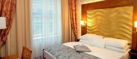 Quarto de hotel com cama, escrivaninha e cadeira. Há uma janela com cortinas, uma parede com papel de parede estampado e uma cabeceira com design exclusivo.