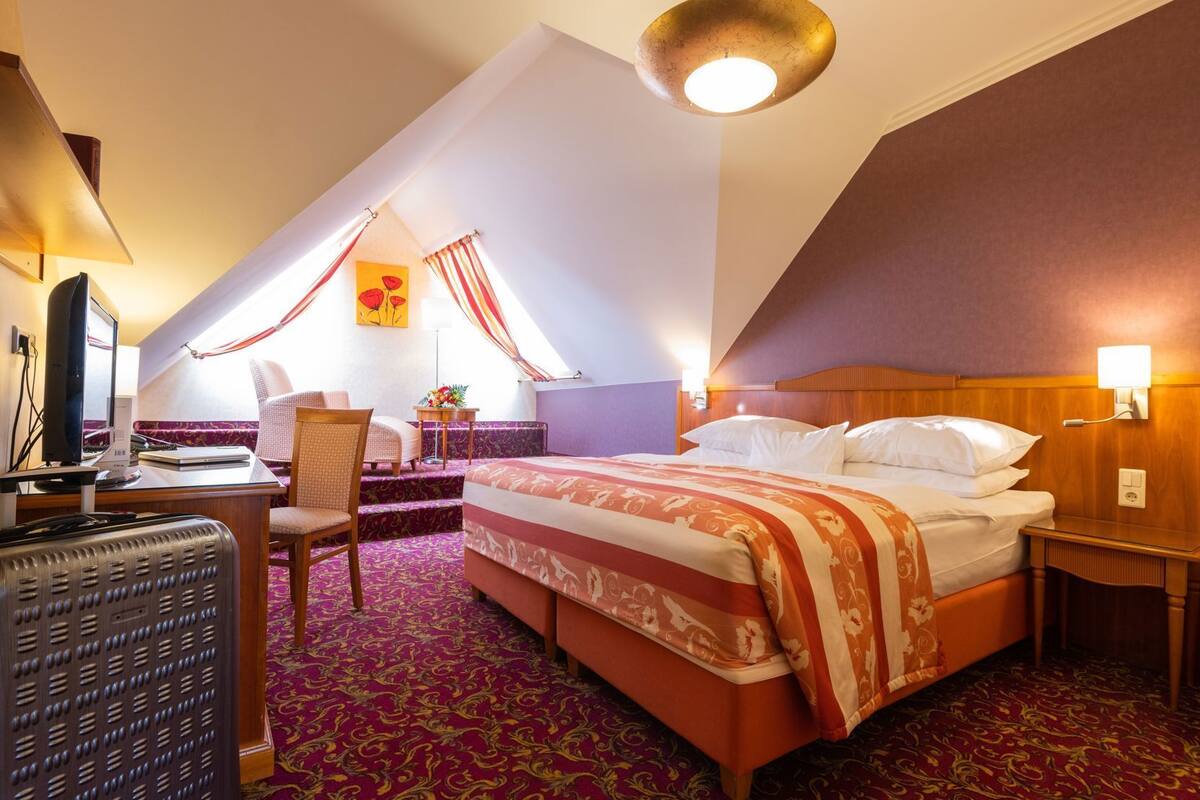 Quarto de hotel com cama, escrivaninha com computador, cadeira, pequena mesa com arranjo de flores e ventilador de teto.