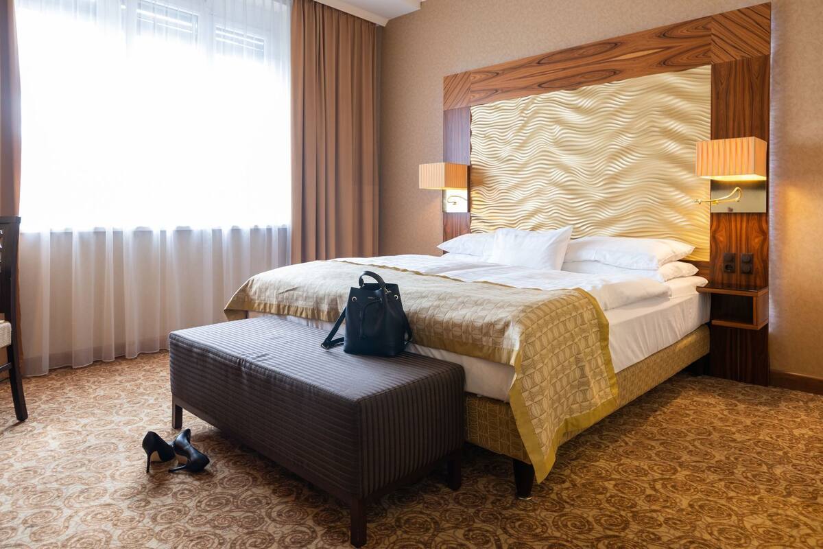 Quarto de hotel com cama, mesa de cabeceira, abajur, cadeira e janela com cortinas.