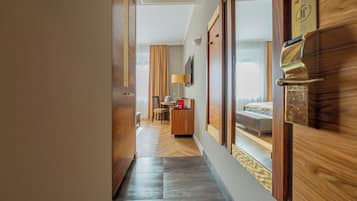 A hotel corridor with a wooden floor, a desk with a lamp, a bed, and a TV on the wall.