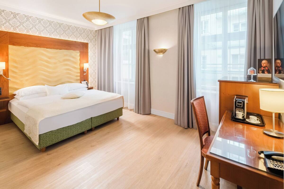 Quarto de hotel com uma cama grande, uma mesa com telefone e cafeteira, e vista para a cidade através de amplas janelas.